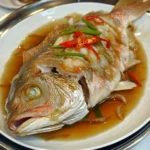 A plate of freshly steamed Chinese fish garnished with ginger, scallions, and red chili slices, served with a drizzle of soy sauce.